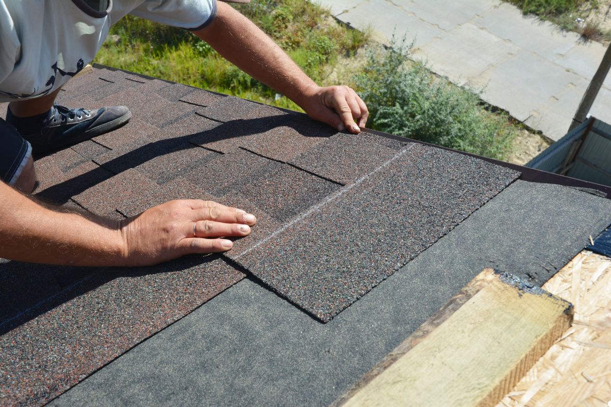 A man carefully installs a roof shingle on a house, using tools and standing on a ladder for support.