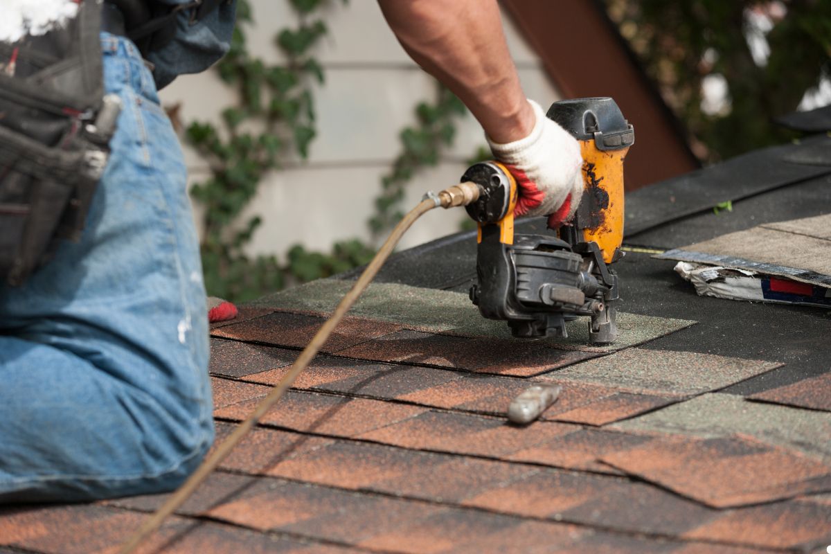 A man operates a nailer to fix shingles on a roof, demonstrating skilled craftsmanship in a sunny outdoor setting.